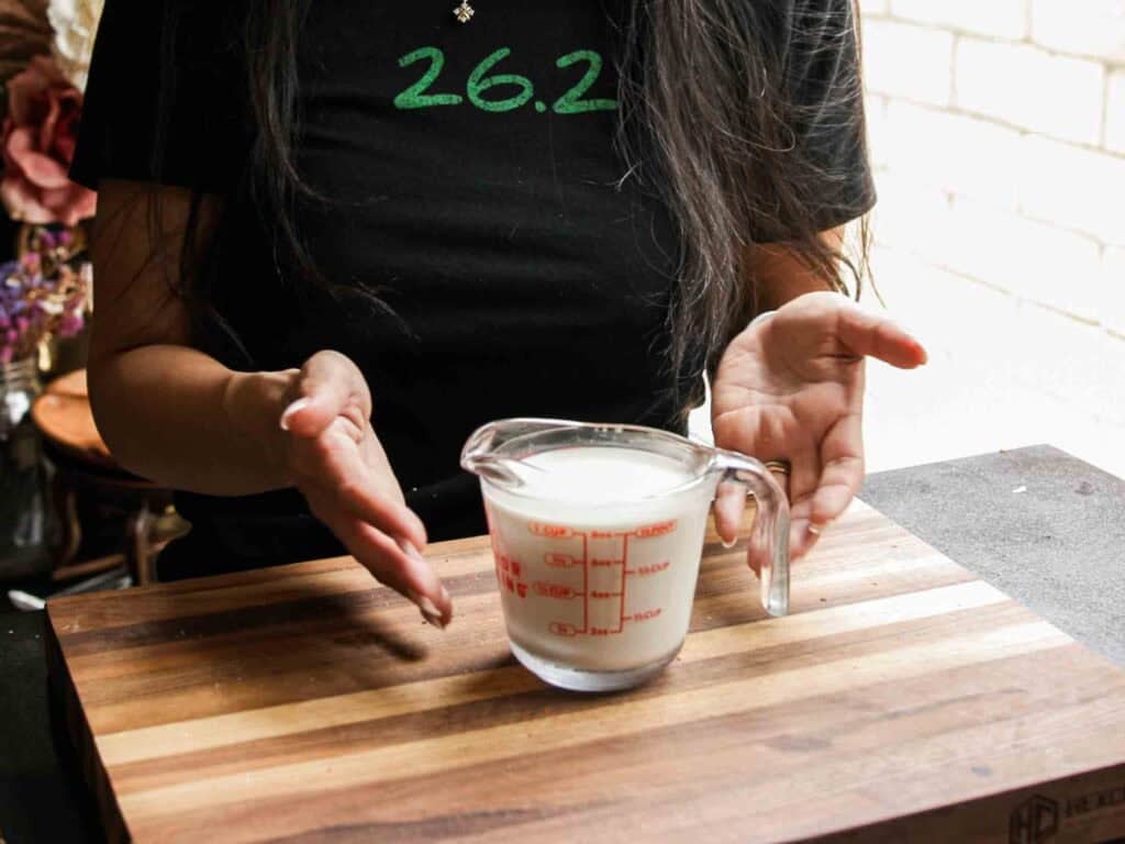 A person with long dark hair gestures toward a glass measuring cup filled with non-dairy milk, placed on a wooden cutting board. The person is wearing a black shirt with “26.2” in green text.
